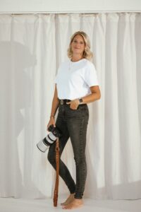 Blonde photographer holding a camera, wearing black jeans and a white t-shirt.