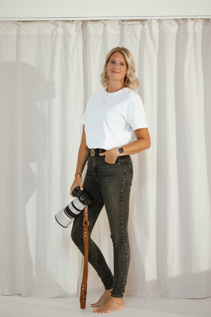 Blonde photographer holding a camera, wearing black jeans and a white t-shirt.